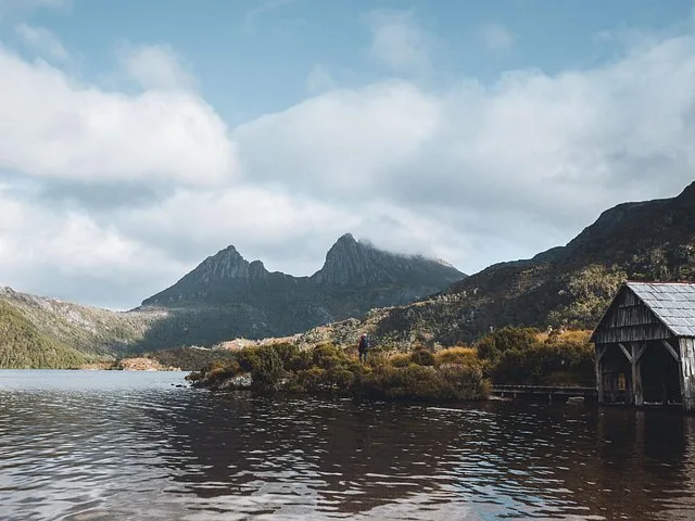 A wooden boatshed by a lake with craggy mountains in the distance.