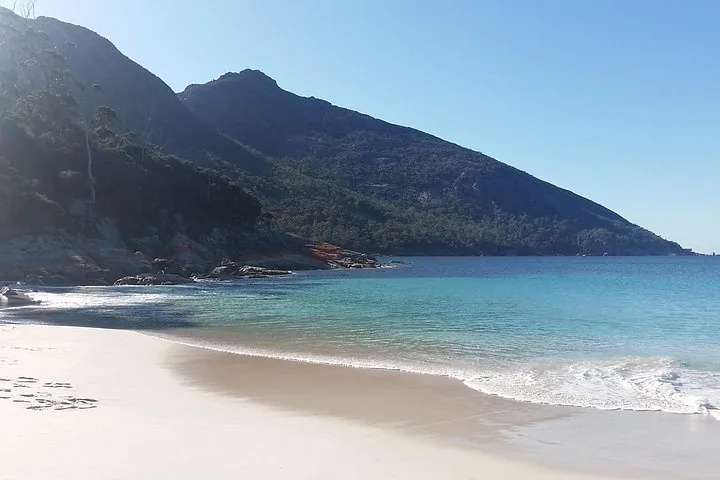 Empty beach with clear blue water and a forested mountain.