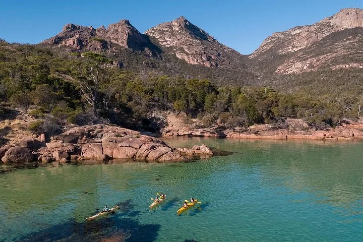 People kayaking in clear water near a rocky shore with mountains behind