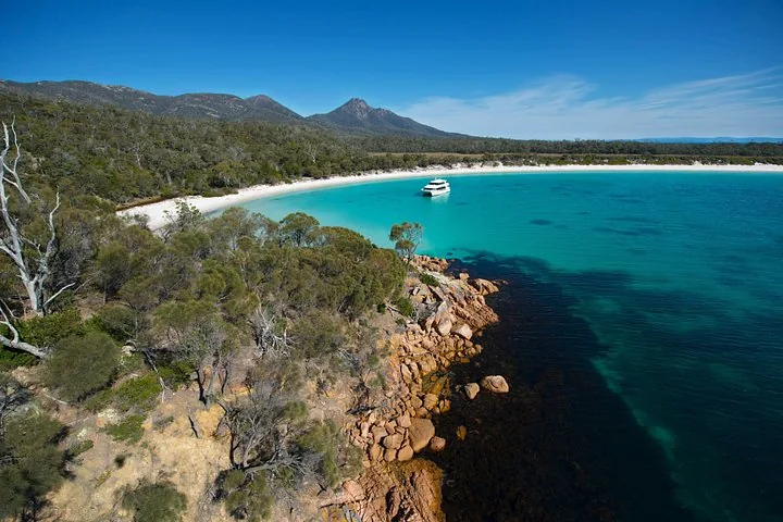 A boat in a turquoise bay surrounded by a sandy beach and forested hills