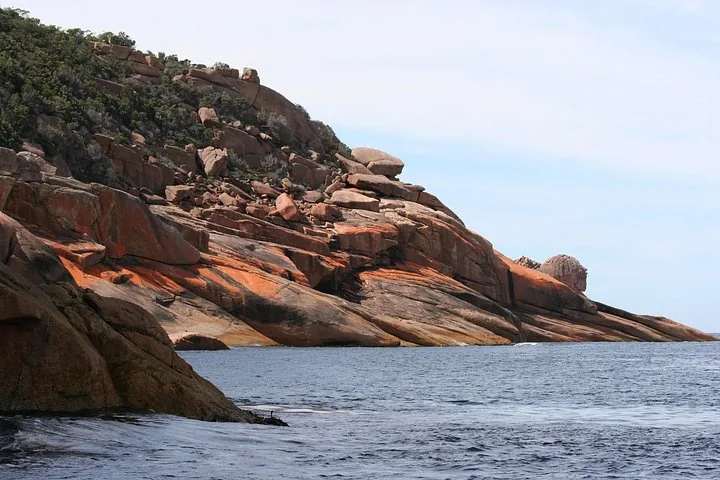 Steep, rocky orange coastline meeting the dark blue ocean water under a partly cloudy sky